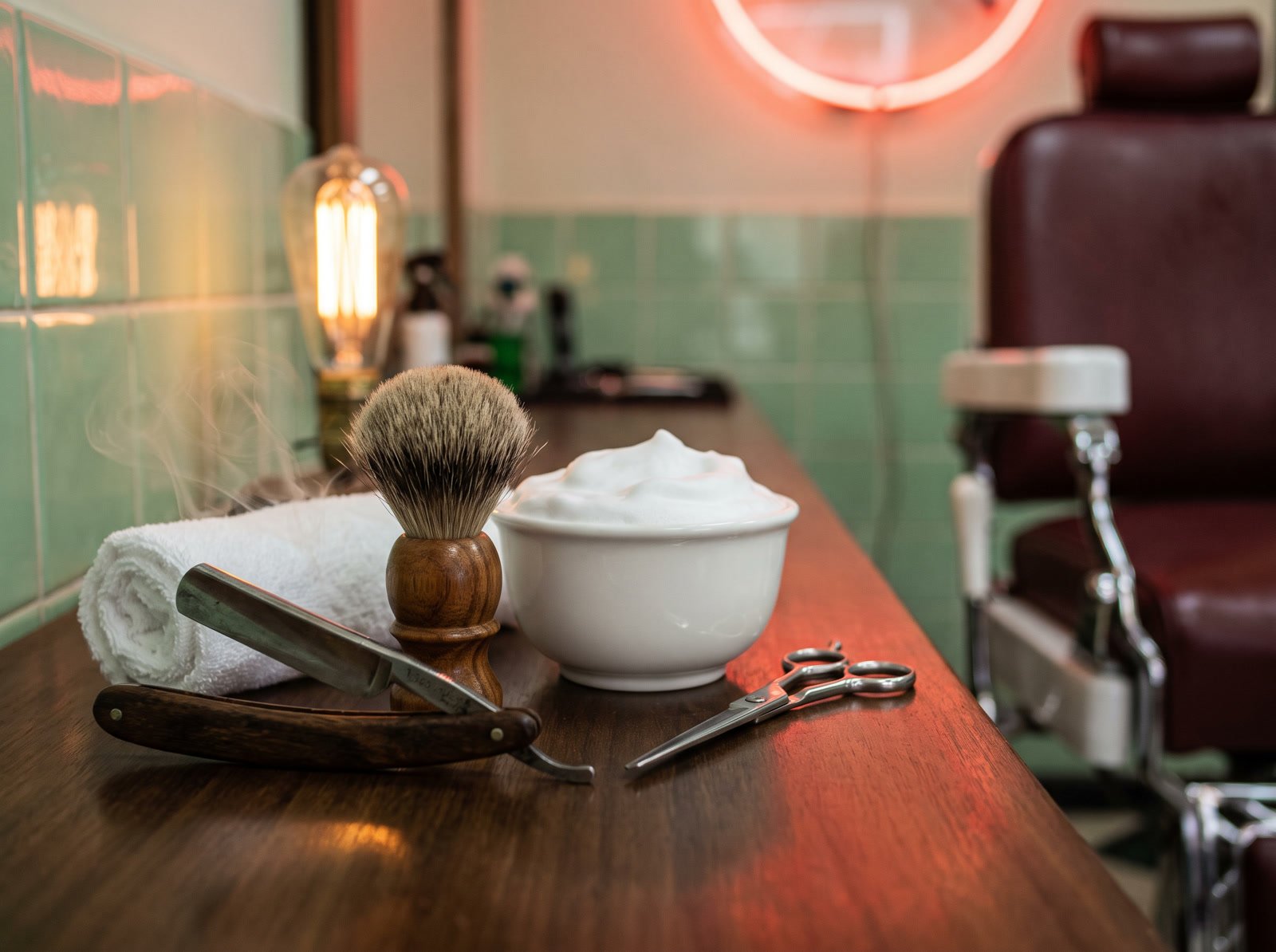 Bancada de madeira com navalha, pincel de barba, cuia de espuma e toalha quente soltando vapor